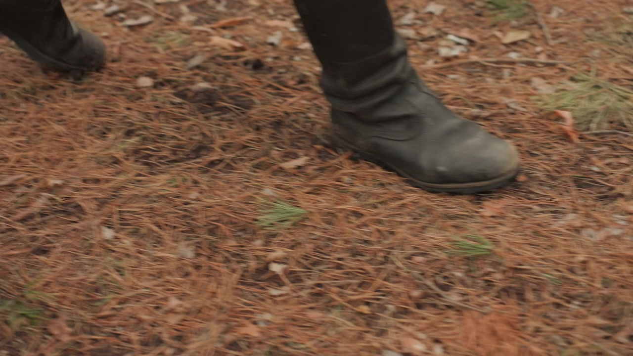 Primer plano en ángulo bajo de piernas y botines oscuros moviéndose sobre el suelo del bosque cubierto de agujas de pino, con una bota apoyada y la otra avanzando, mostrando un movimiento natural y constante al caminar.