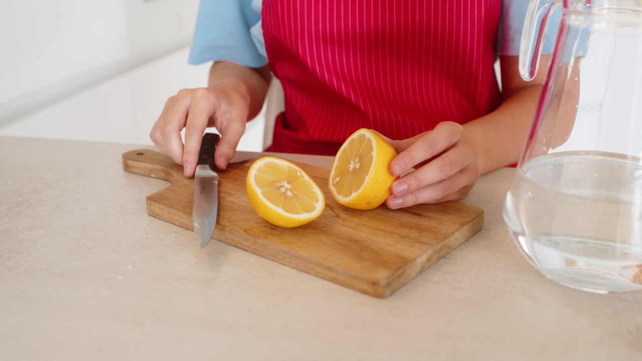 Young woman in kitchen wearing apron cuts lemon on wooden board next to water jug clean fresh prep