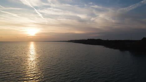 Aerial-reverse-dolly-above-coastal-region-at-sunset,-golden-hour-ray-on-water