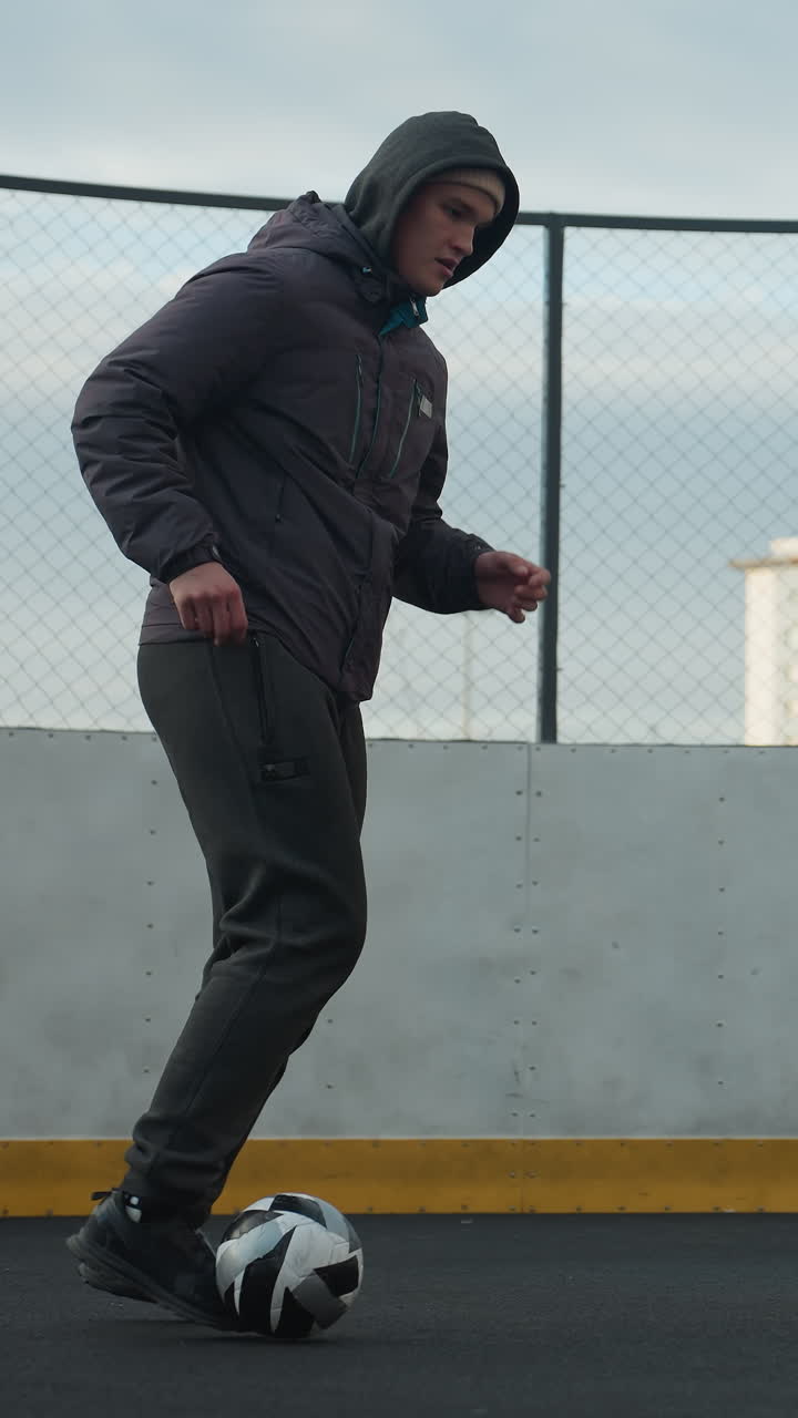 hombre practicando ejercicios de fútbol en la arena deportiva, empujando hábilmente la pelota mientras parece cansado, poste de la portería y edificios residenciales urbanos en el fondo