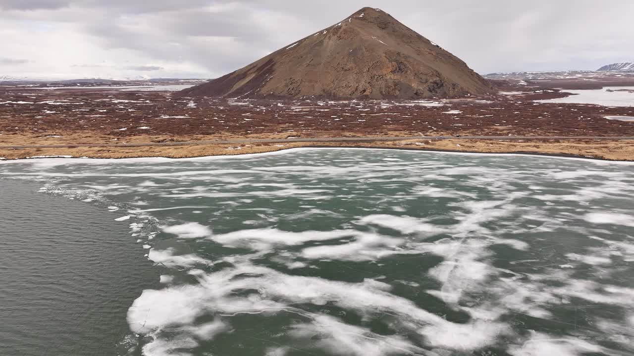 Aerial view of frozen shoreline meeting open water with volcanic hill near Skútustaðir. Reykjahlíð, Iceland