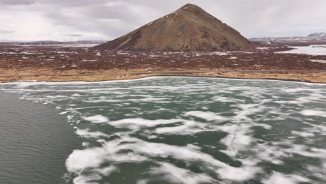 aerial view of frozen shoreline meeting open water with volcanic hill near skútustaðir. reykjahlíð, iceland