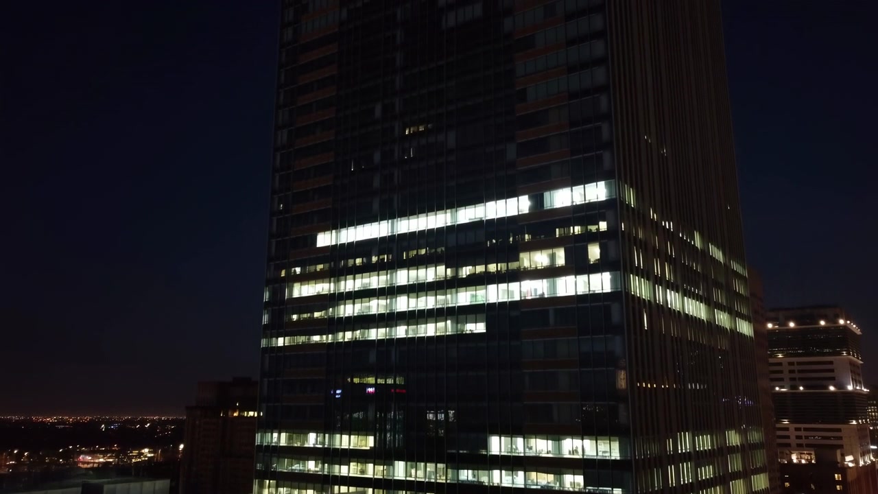 Arranha-céu de escritórios de vidro alto visto do nível da rua para cima à noite, com iluminação interior clareando progressivamente nos andares inferior, médio e superior, criando faixas quentes e brilhantes contra.