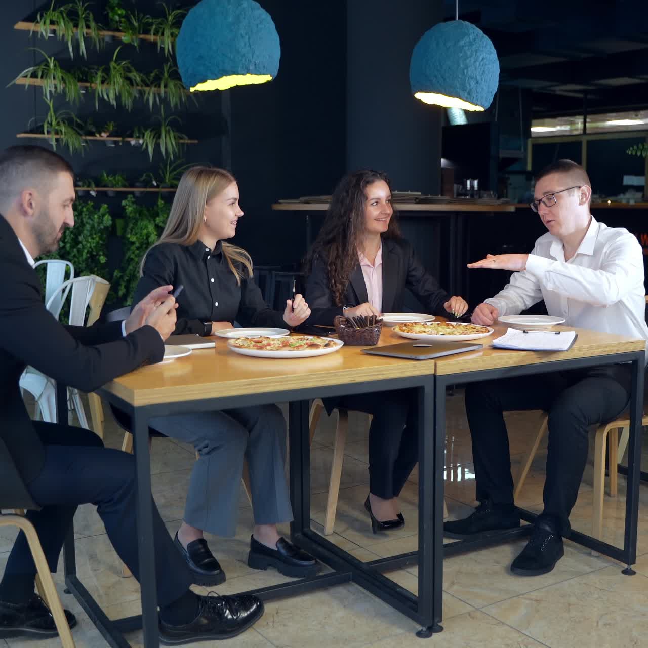 Teammates sitting in a buffet at lunchtime. Man and two women speaking and gesturing. Other man is concentrated on his smartphone
