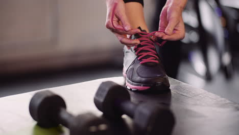 low section of female athlete tying lace of sports shoe by dumbbells at fitness studio