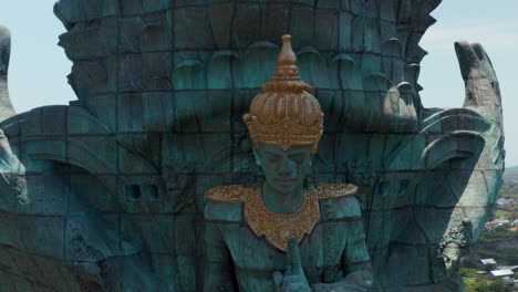 Close-up-view-of-Garuda-Wisnu-Kencana-statue-in-park-in-Bali,-Indonesia,-rising-above-the-city.-Aerial-retreating-reveal-of-large-blue-and-green-statue-of-Hindu-religion