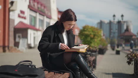 young lady seated outdoors on a bench reading her book, with a focus on the book's pages, she is wearing black boots and a coat while a bag rests beside her, urban setting in the background