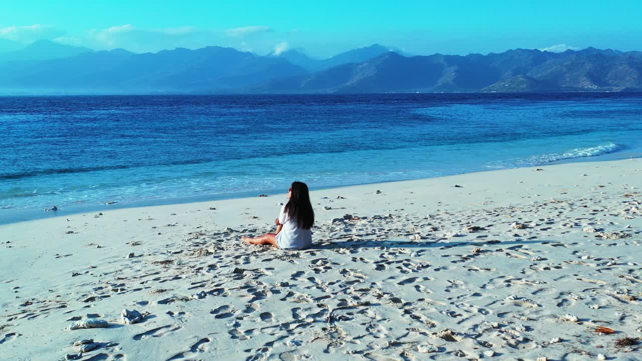 Tourist girl in white t-shirt sitting on shining sand at sunrise, watching calm blue sea and mountains horizon under bright sky in Indonesia