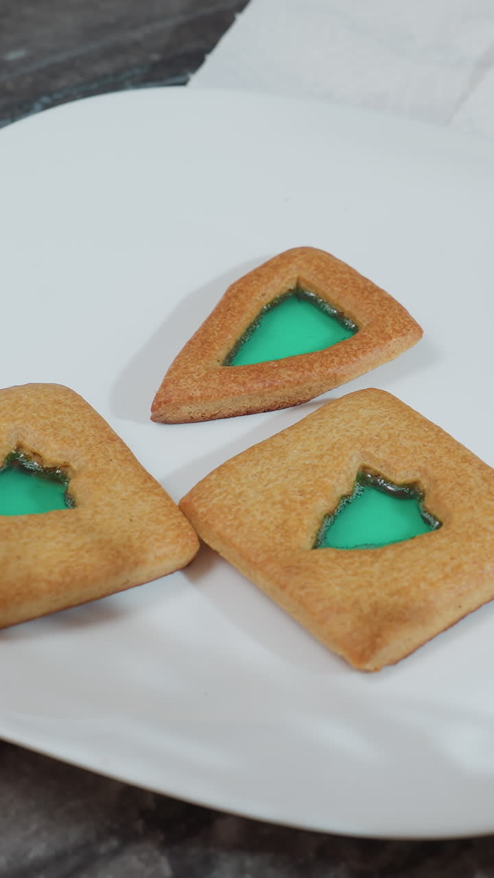 Three decorated cookies on white plate with green filling, red burning candle, and decorative flower on table, two other cookies placed on white textured paper