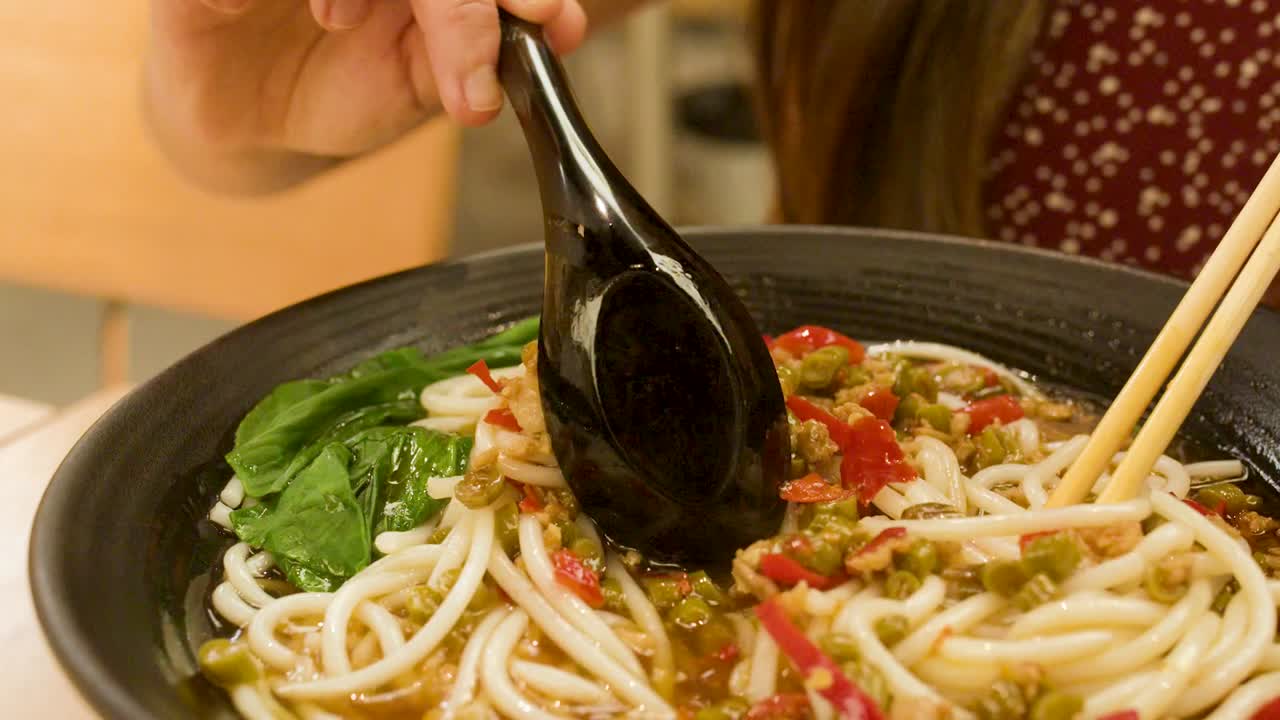 Una donna usa un cucchiaio nero e delle bacchette per mescolare noodles piccanti e verdure