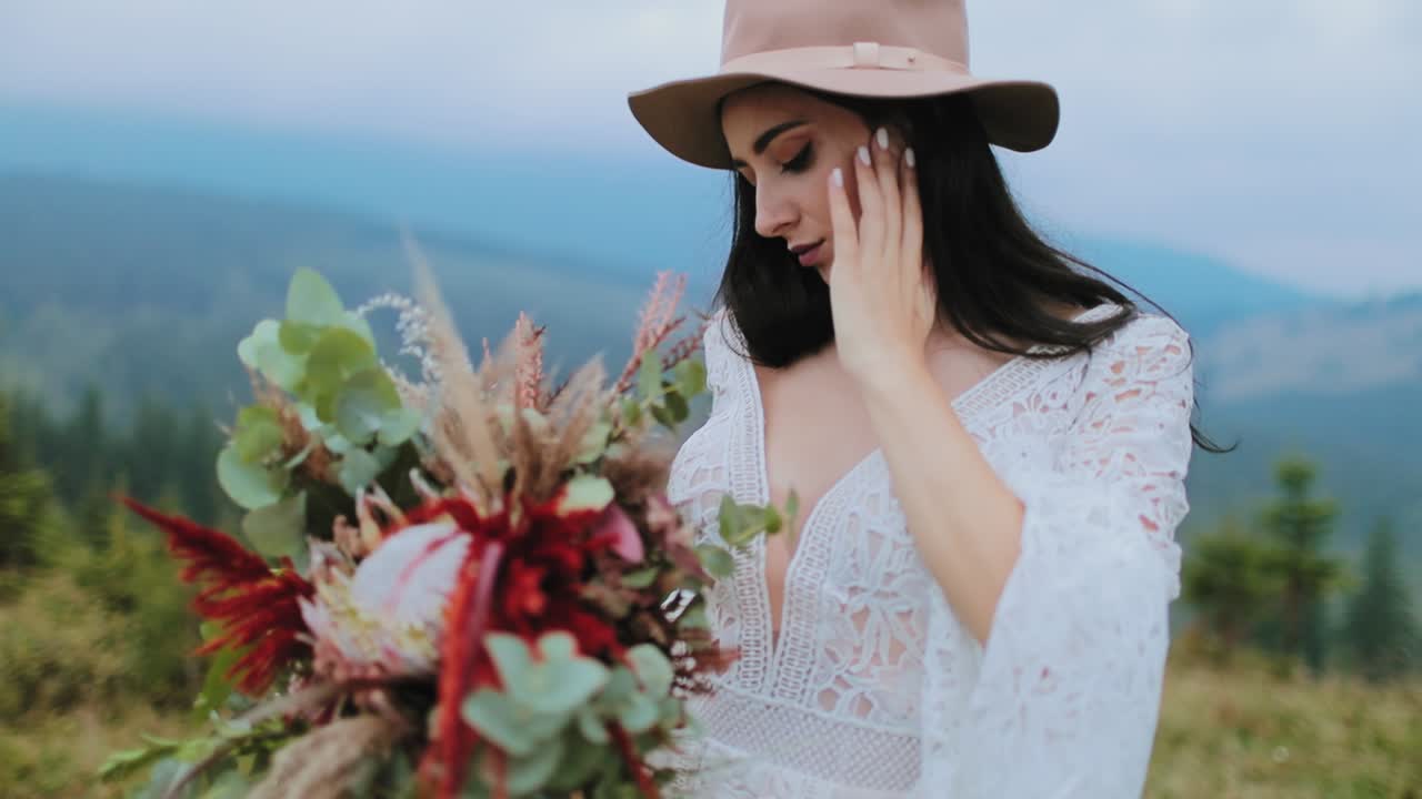 Lovely woman in hat and white dress in nature. Beautiful model with dark hair holding bouquet of flowers and posing to camera.