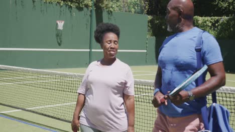Glückliches-älteres-Afroamerikanisches-Paar,-Das-Mit-Tennisschlägern-Spaziert-Und-Sich-Auf-Dem-Tennisplatz-Unterhält