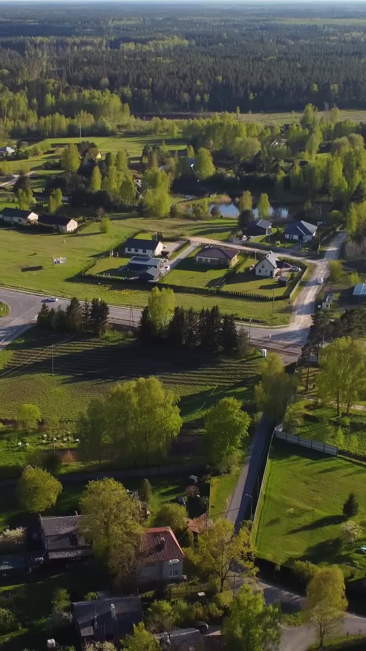 Vertical drone pull away reveals Katlakalns spring residential area with green fields, country roads, traffic, and houses surrounded by trees under clear sky and golden afternoon light