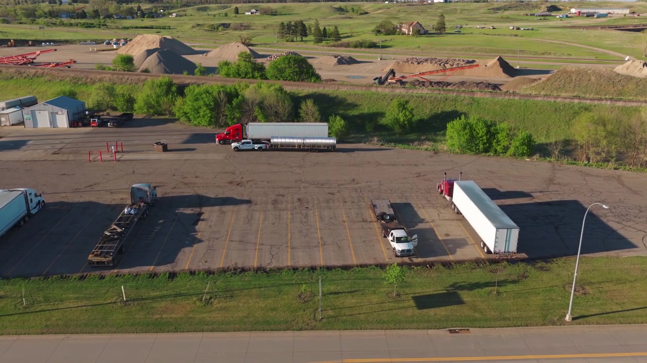 Aerial truck stop at Jamestown ND — semi trailers, tanker and flatbed parked near a gravel yard and rail line, trucking industry, spring evening.