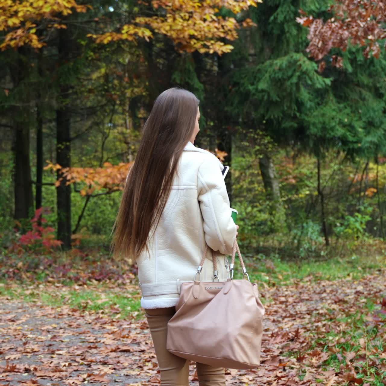 Giovane donna alla moda con una grande borsa che passeggia nel parco autunnale. Vista posteriore. La donna si volta verso la fotocamera tenendo in mano un telefono e un bicchiere di carta