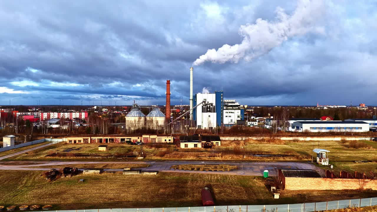 A Factory Complex Releases a Plume of Smoke Into the Sky, Standing Against a Dramatic Backdrop of Dark Clouds and Open Fields - Wide Shot