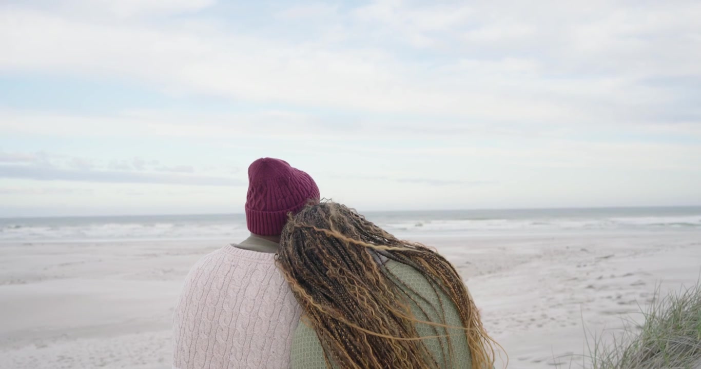 Afroamerikansk par sidder på stranden, mand med hue taler, kvinde læner sig og ser på havet