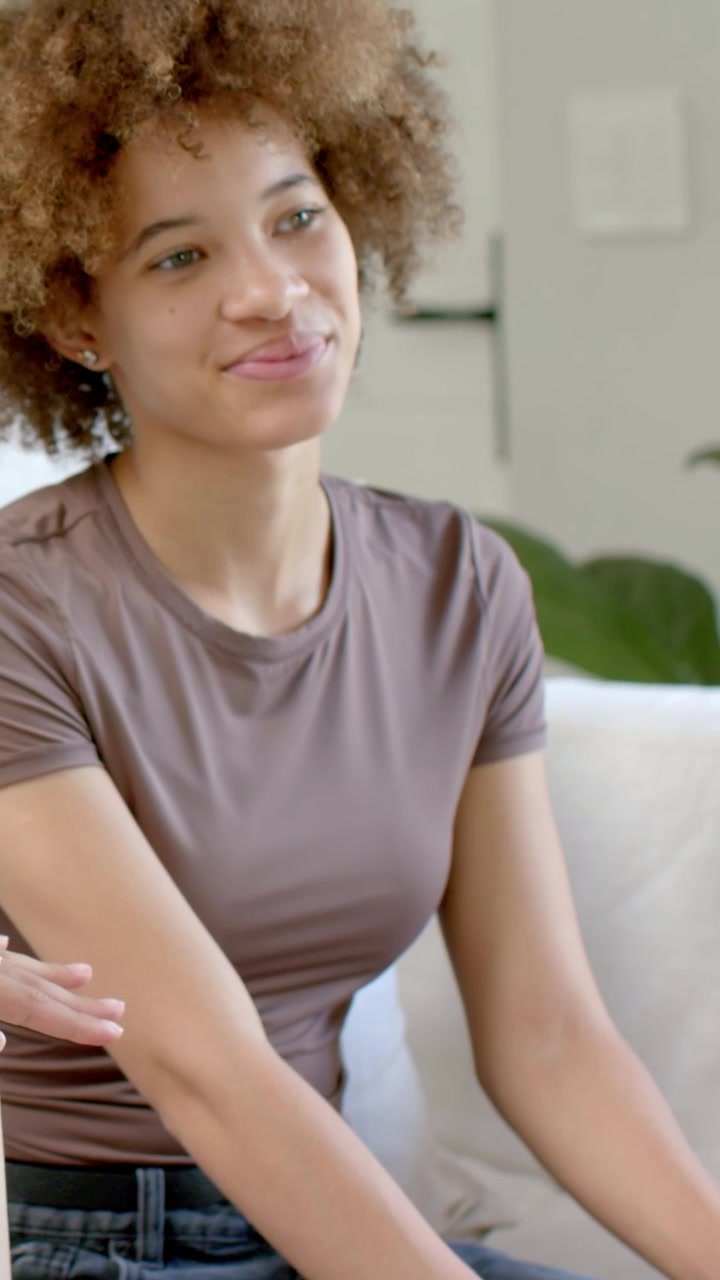Vertical video: Smiling woman in brown tee on sofa listening in chat while hands placing beige box