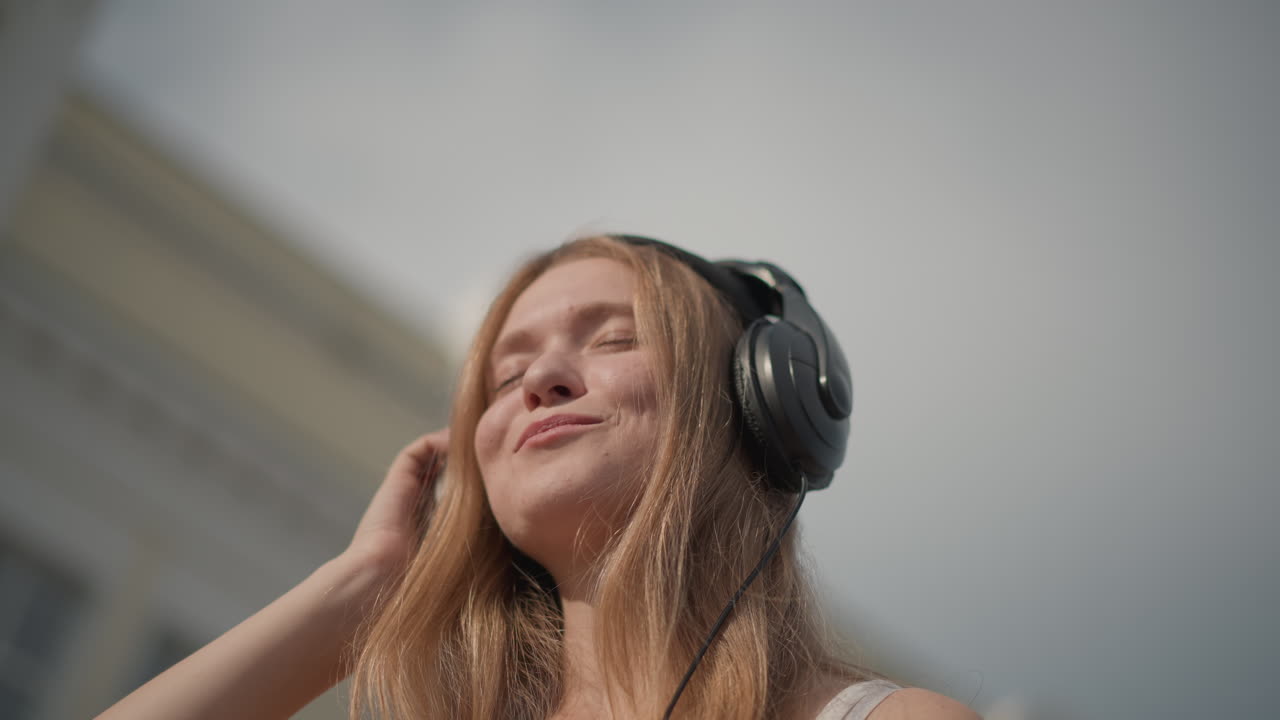 Donna caucasica in strada soleggiata con cuffie, occhi chiusi e sorridente mentre si sistema il berretto, capelli lunghi che catturano la luce del sole e le particelle di polvere, primi piani che catturano uno stato d'animo introspettivo, testa delicata
