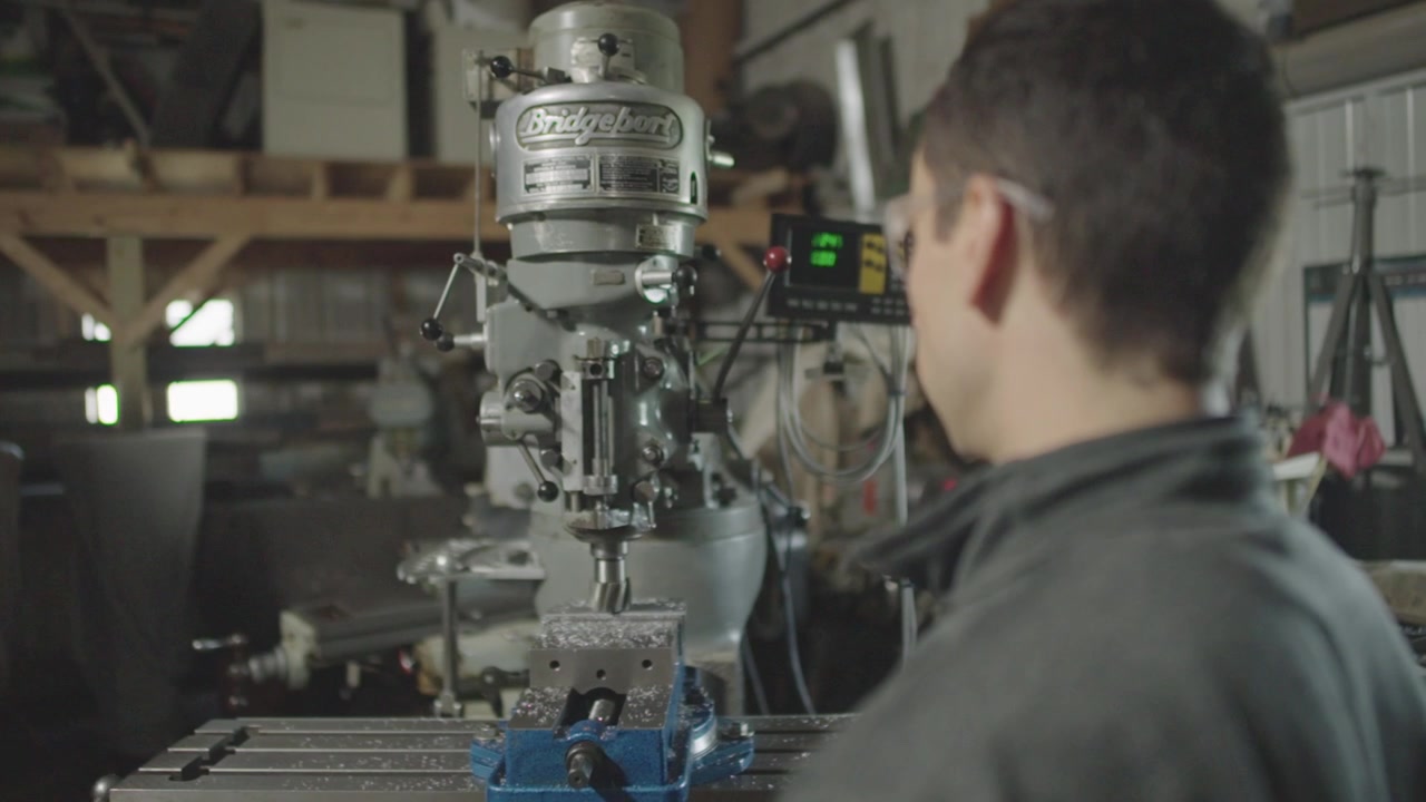 An over-the-shoulder, slow motion shot of a young mechanic overseeing their old Bridgeport machine with a high speed drill in their workshop