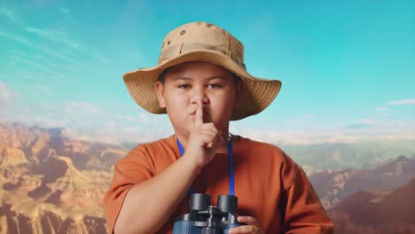 asian boy with a hat looking through binoculars then making shh gesture while traveling at the top of mountain. boy researcher examines something, travel tourism adventure concept, close up