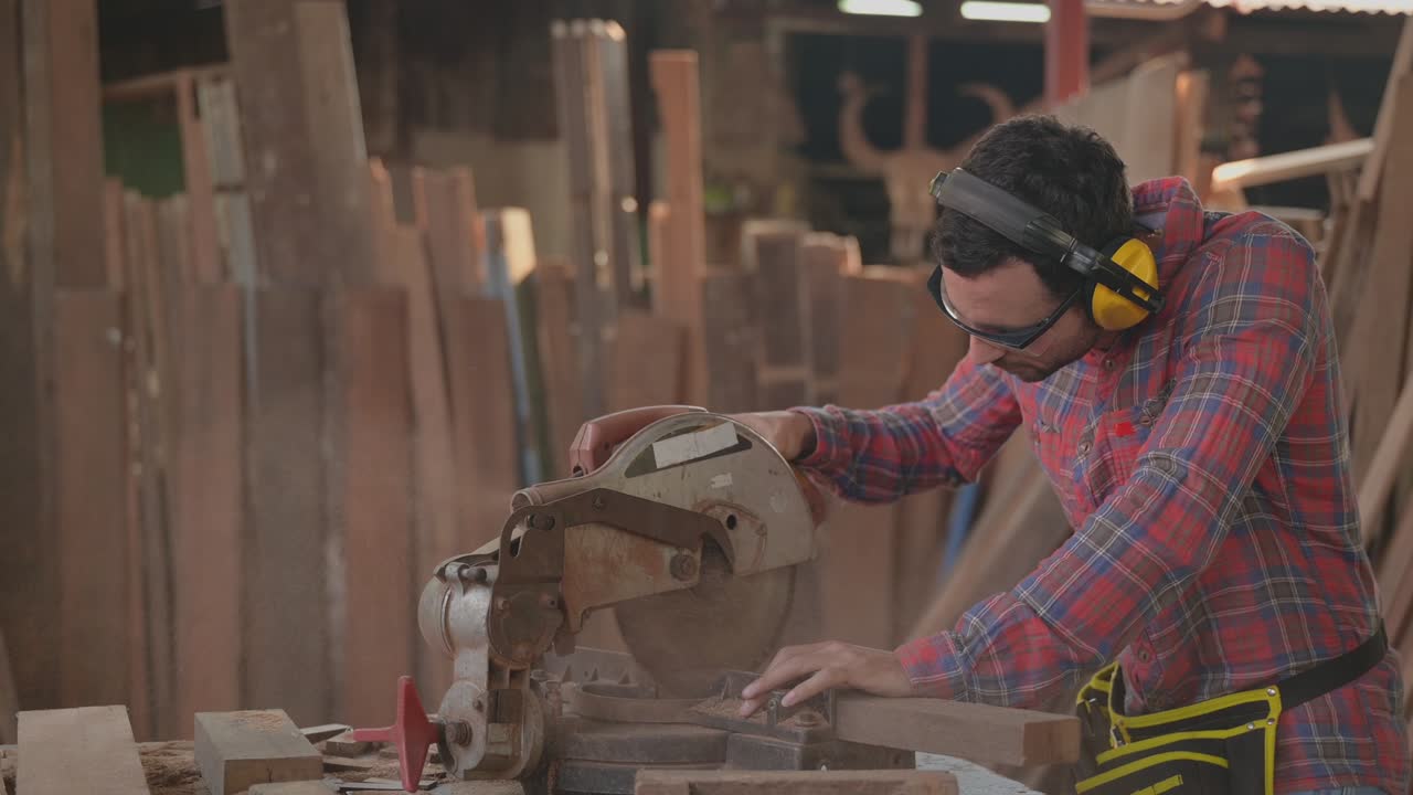 красивый плотник, носящий очки и наушники для личной безопасности, использующий круговую пилу для мебели на рабочем месте на заводе.