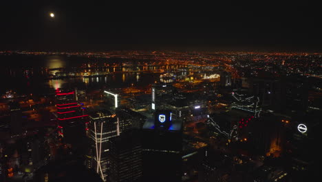 Illuminated-modern-high-rise-downtown-buildings-and-harbour-on-coast.-Backwards-fly-above-night-metropolis.-Moonshine-reflection-on-water-surface.-Cape-Town,-South-Africa