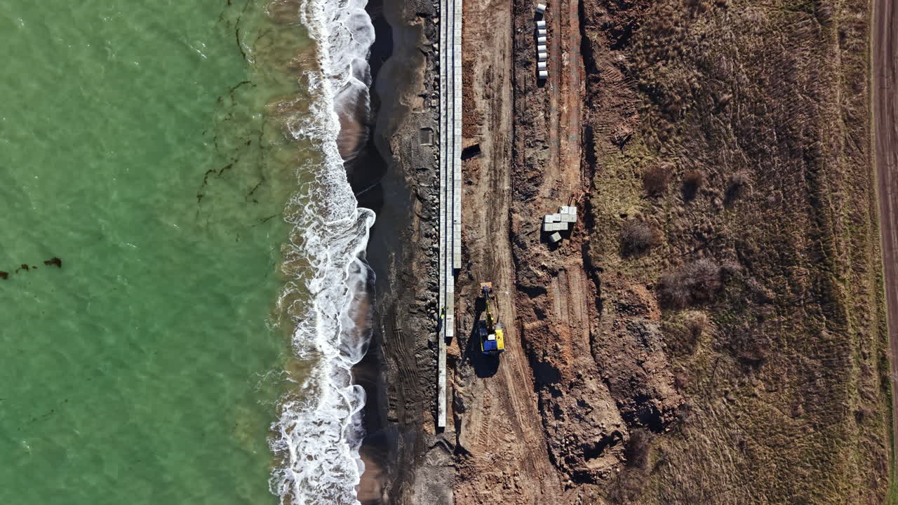 Lavori di costruzione in corso vicino alla riva, mentre macchinari e materiali vengono disposti per un progetto lungo la costa durante il giorno.