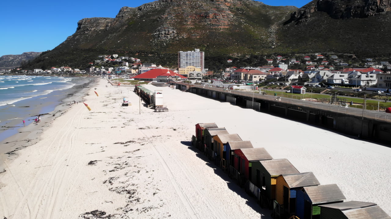 Aerial View Sandy Beach Huts Lined Along White Shoreline Under Mountain Backdrop, Quiet Coastal Town. Media