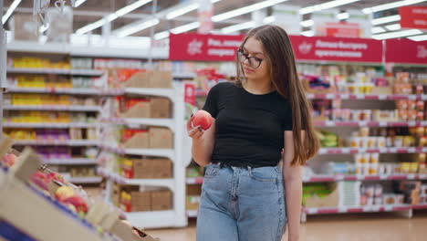 young woman in grocery shop picks red apple from display, observing it, and drops it back, price tags and signposts visible in bright, vibrant store setting