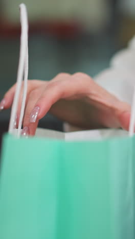 primer plano de una mano con uñas pulidas abriendo con gracia una bolsa de compras de color menta mientras está sentada, las vibrantes bolsas verdes y rosas mejoran la elegante escena de compras