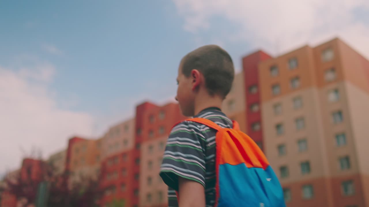 niño pequeño caminando por la calle con mochila en una zona urbana industrial con casas de paneles en el fondo