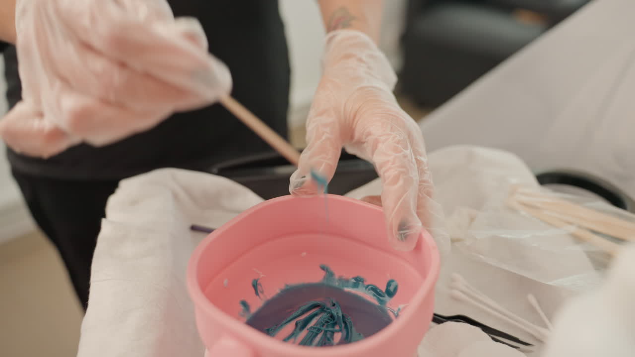 Mani guantate che mescolano una tinta color sarcelt per capelli, ciotola rosa con un bastoncino di legno su un asciugamano nella postazione del salone, colorista che prepara una formula cremosa di pigmenti per l'applicazione, primo piano che mostra guanti igienici e precisione