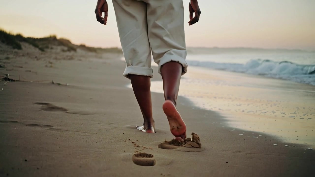 Donna adulta che cammina lasciando impronte a piedi nudi nella sabbia bagnata lungo la riva, con i pantaloni arrotolati, all'alba.
