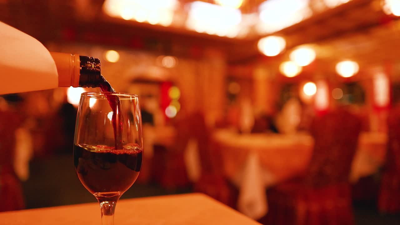 A hand pours red wine into a glass on a table in a warmly lit, upscale restaurant with blurred background and soft ambient lighting