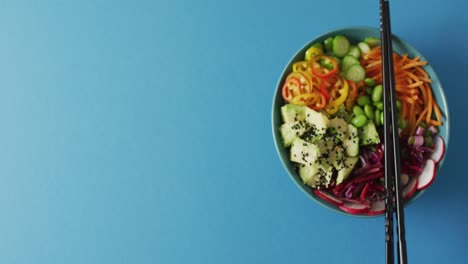 Composition-of-bowl-of-rice-and-vegetables-with-chopsticks-on-blue-background