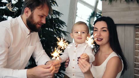 familia feliz y fuego de bengala en casa junto al árbol de navidad. madre y padre sosteniendo bengala, mientras su lindo hijo está