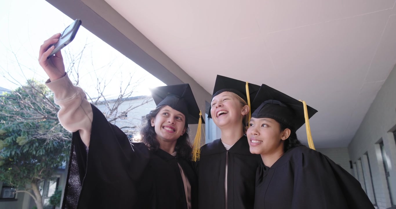 三名女青少年学生在毕业典礼上戴着学士帽，举起手机自拍并查看照片。