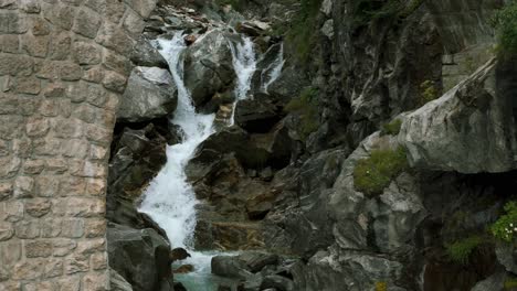 Fresh-water-of-mountain-waterfall-of-Furka-Pass-in-Switzerland-flowing-toward-brick-bridge