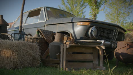 Coche-Viejo-Y-Oxidado,-Plano-Amplio-Y-Bajo,-Rodeado-De-Herramientas-Agrícolas-Rústicas