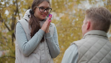Rear view of man on knees proposing to surprised woman outdoors as she nods in agreement with emotional smile while wind sways autumn leaves around romantic couple in matching outfits
