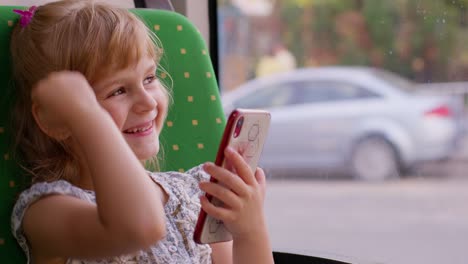 smiling child girl in city bus or tram transport use mobile cell phone typing browsing say wow yes