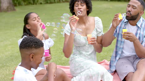 Felices-Padres-Afroamericanos,-Hija-E-Hijo-Soplando-Burbujas-Sentados-En-El-Jardín,-Cámara-Lenta