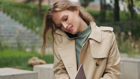 Caucasian-female-student-talking-on-the-phone-while-writing-in-a-notebook-outdoors.