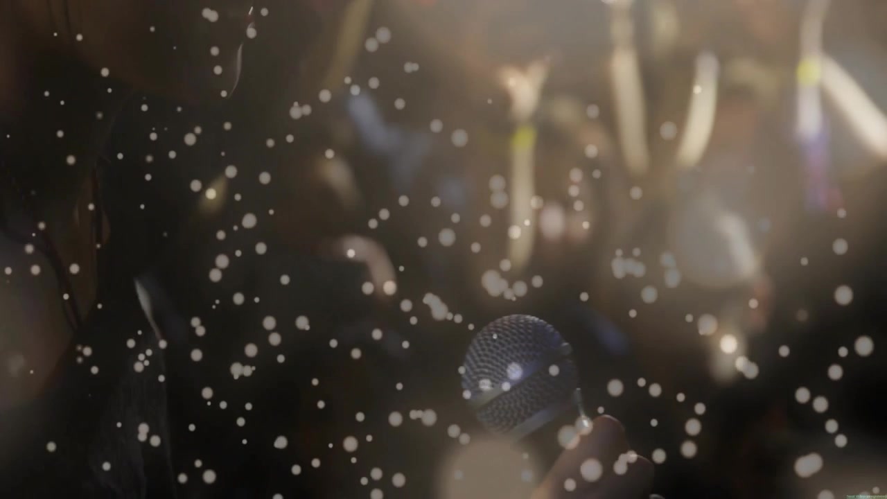 Kamera menarik fokus, penyanyi wanita muncul di antara bokeh dan konfeti dengan mikrofon, menampilkan musik.