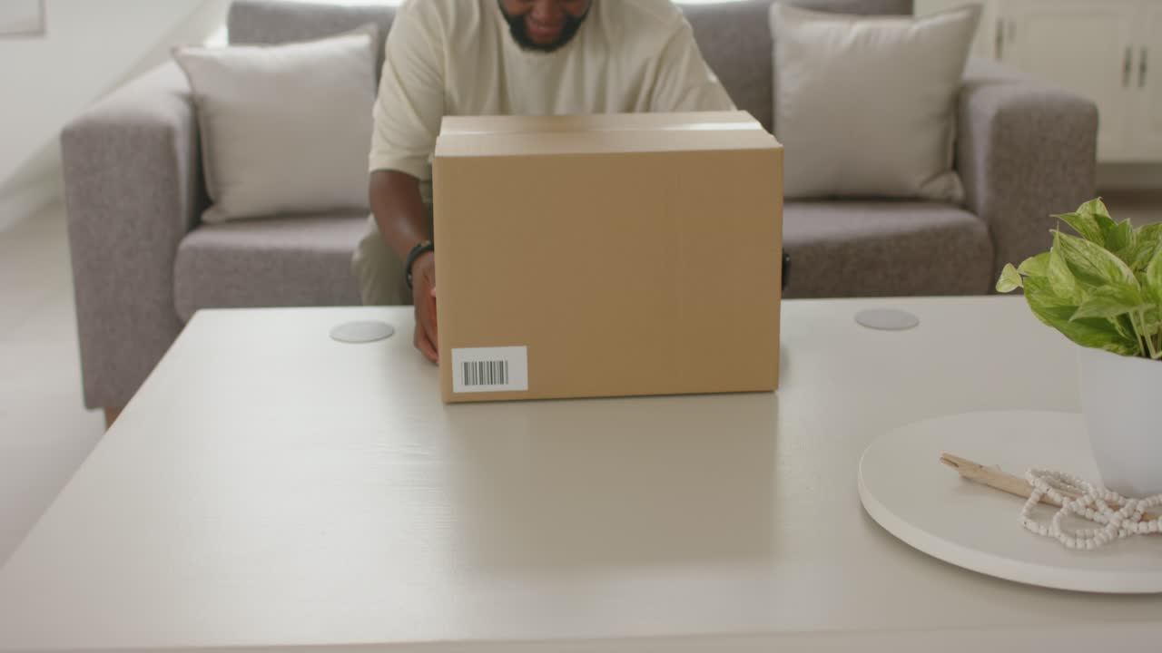 Hombre afroamericano cargando una caja después de la entrega, colocándola en una mesa de centro blanca, preparándose para abrirla.