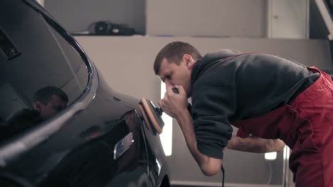 un joven trabajador de un centro automotriz cuida un coche caro negro, puliéndolo con una máquina de frotar.