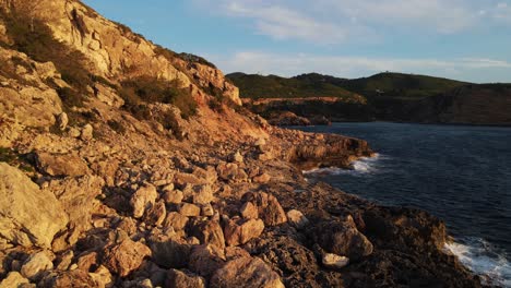 Aerial-view-coastal-rocky-cliffs-of-bay-of-Cala-Xarraca-in-Ibiza,-Spain-as-waves-crash-on-the-rocks-with-mountains-and-blue-sky-in-the-background