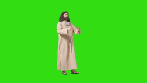 studio shot of man wearing robes and sandals with long hair and beard representing figure of jesus christ raising arms and praying on green screen