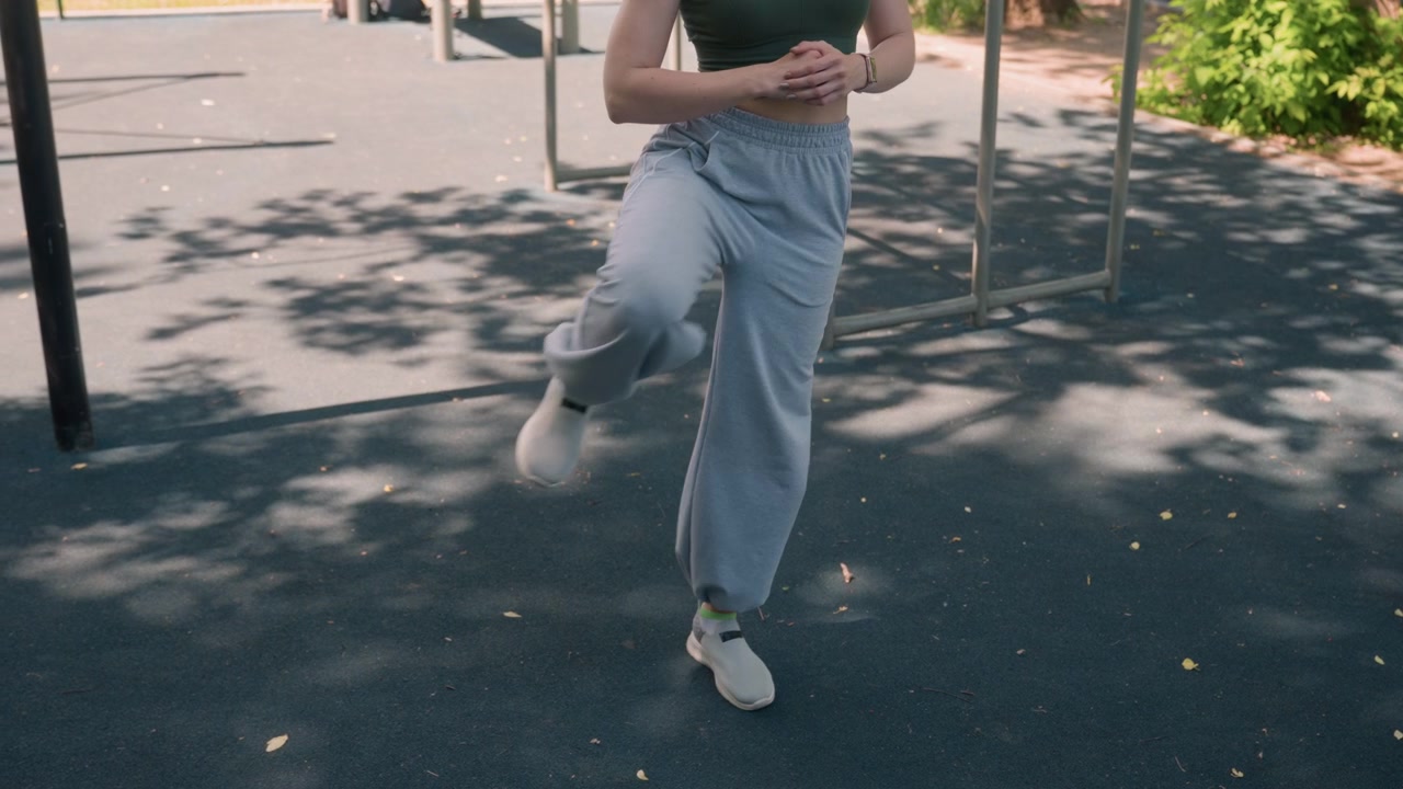 Primo piano di una giovane donna che esegue esercizi di sollevamento delle ginocchia in un parco all'aperto. La donna indossa abbigliamento sportivo casual e muove le gambe ritmicamente con la luce del sole che filtra tra gli alberi.